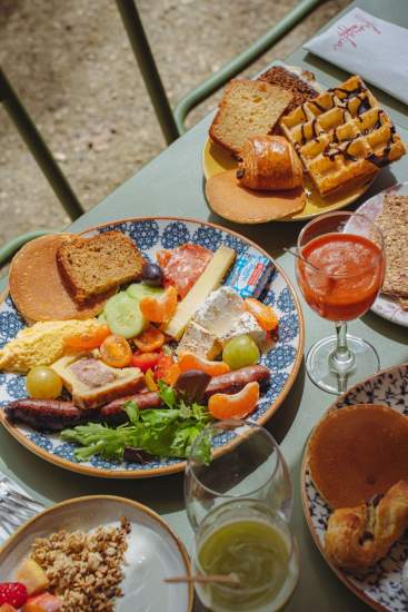 Assiette brunch généreuse avec œufs, saucisse, fromages, légumes frais, pain grillé et gaufre, accompagnée d’un cocktail sur la terrasse de La Cabane Meudon
