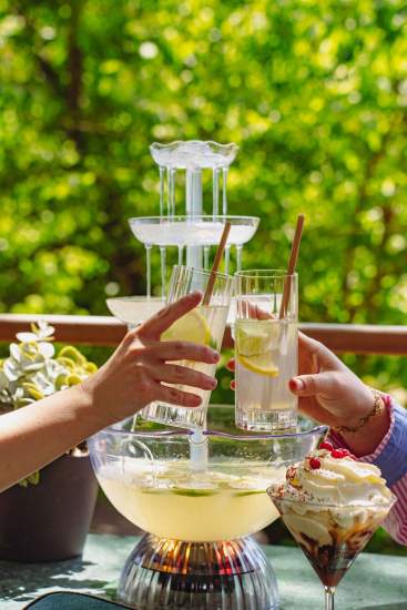 Deux personnes trinquant avec des cocktails rafraîchissants en terrasse, fontaine à boissons en arrière-plan dans un cadre verdoyant à La Cabane Meudon