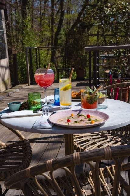 Table en terrasse au soleil avec cocktails colorés et assiette gastronomique, entourée de nature verdoyante